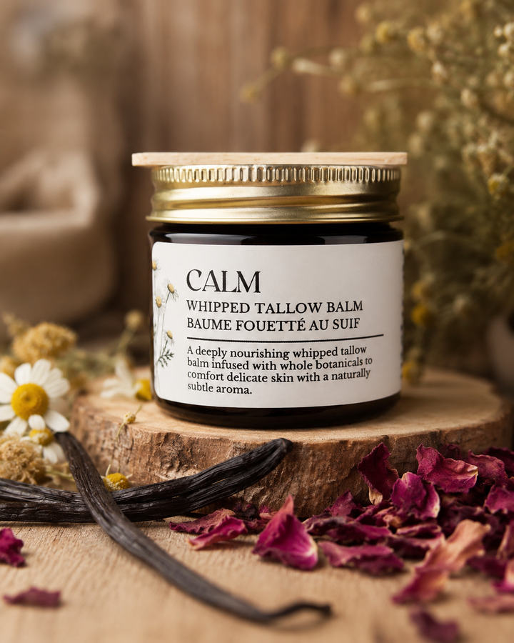 A small glass jar of CALM Whipped Tallow Balm with a gold metal lid and white label, displayed on a rustic wooden slice with dried rose petals, vanilla beans, and daisies in a warm natural setting.