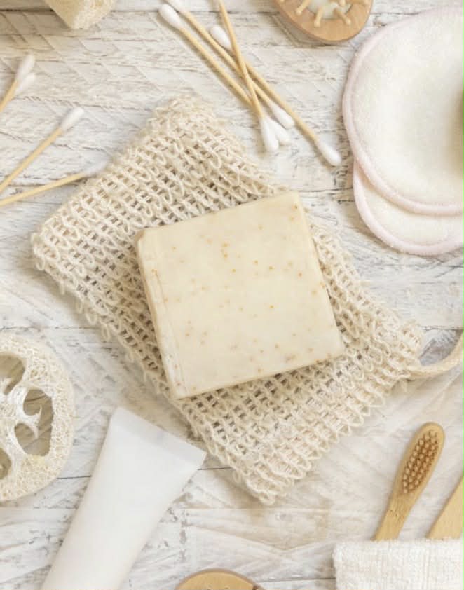 Bar of soap on a natural cotton fiber soap bag with bamboo toothbrushes and loofah on a light wooden surface.