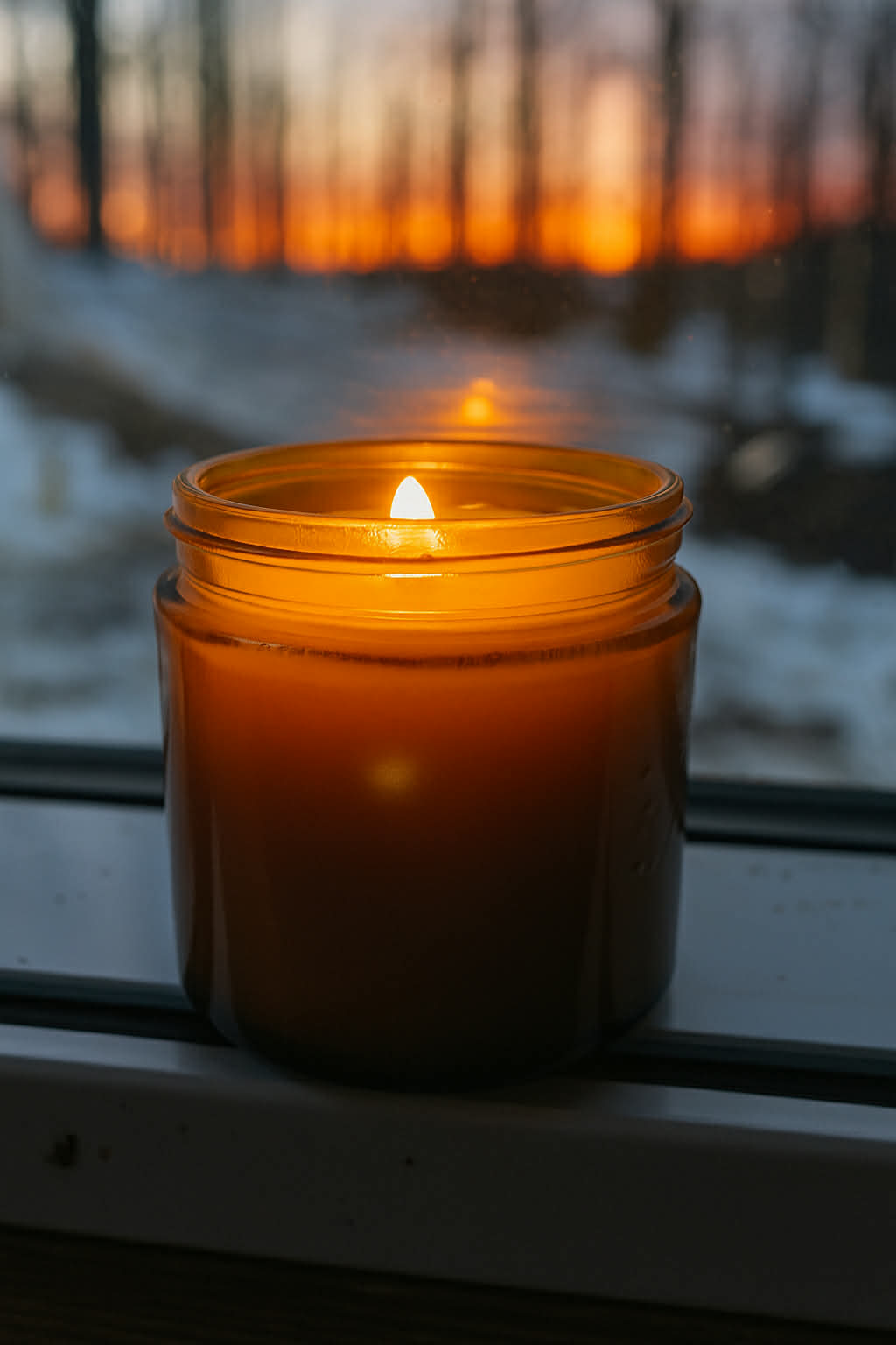 Sage Creek Botanicals lit Candle in an amber jar on a windowsill with a blurred outdoor background