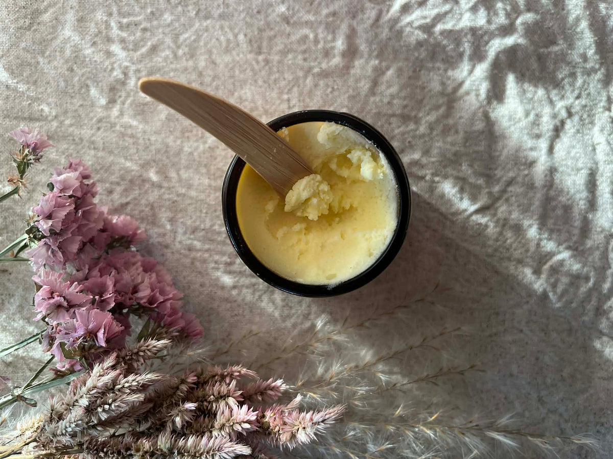 Jar of Sage Creek Botanicals Restorative balm with a wooden spoon on a textured surface with pink flowers.
