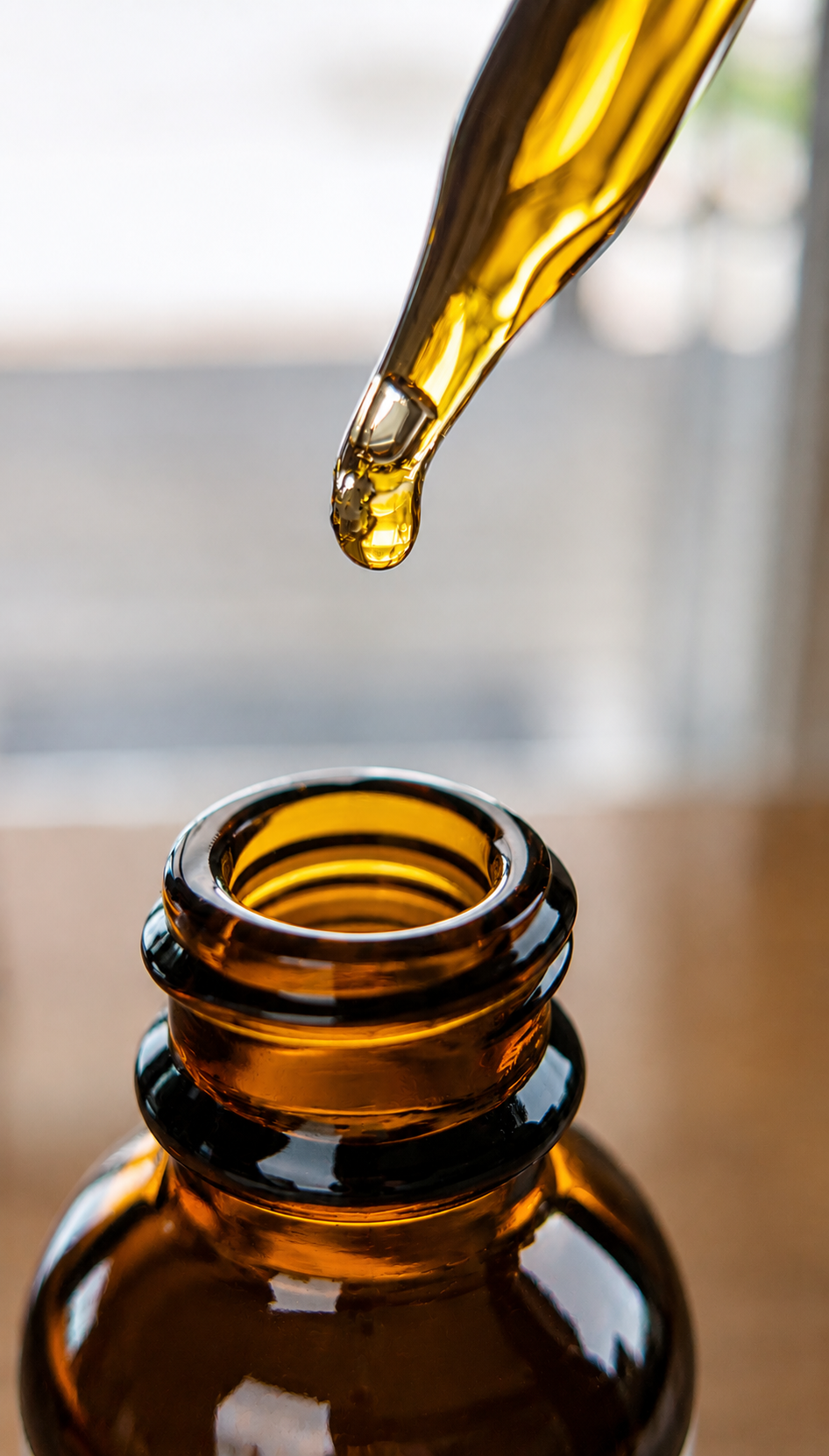 A close-up of an amber glass dropper above an amber glass bottle, with a single drop of golden liquid hanging between them.